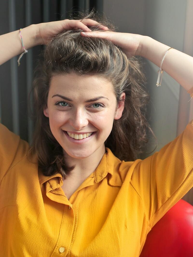 Cheerful woman sitting indoors, smiling brightly with hands behind head, wearing a yellow shirt.