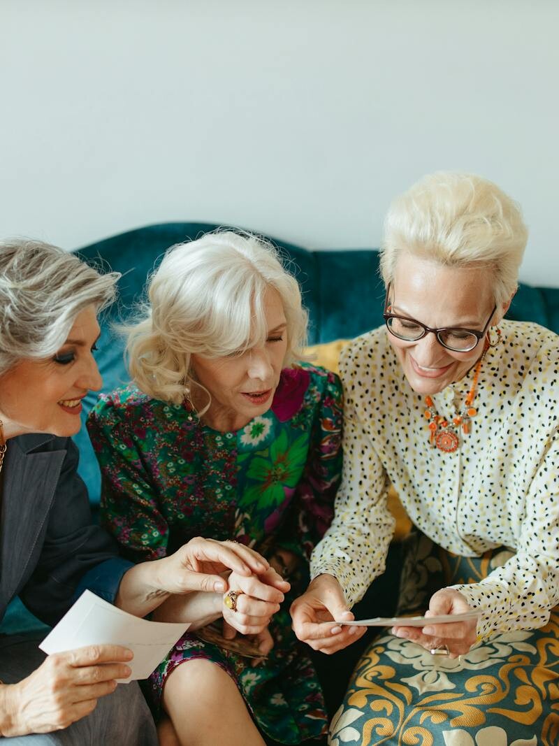 Three senior women joyfully reminiscing over old photos on a cozy couch.