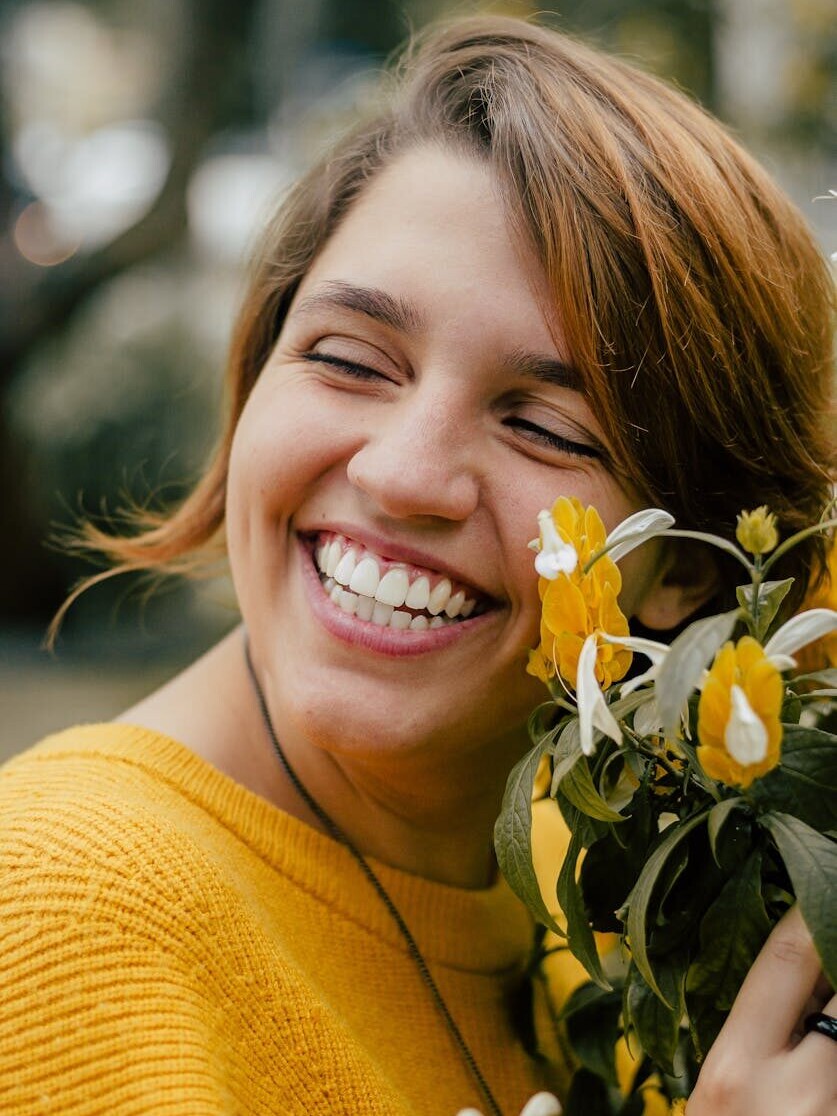 A smiling woman enjoys a sunny day outdoors holding bright yellow flowers.