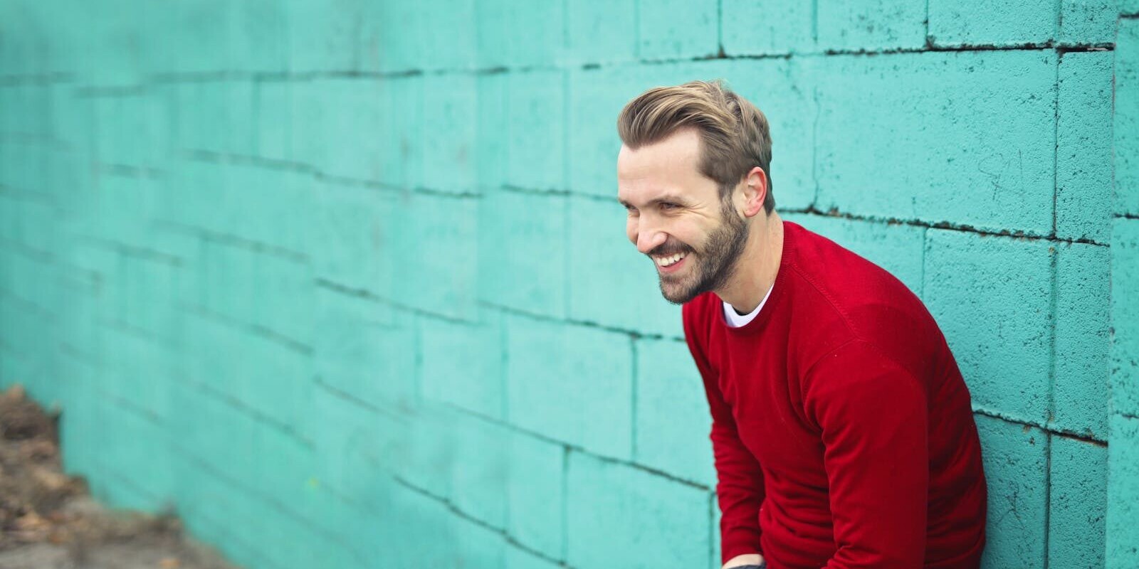 Smiling man in Budapest wearing a red sweater against a teal brick wall, exuding happiness.