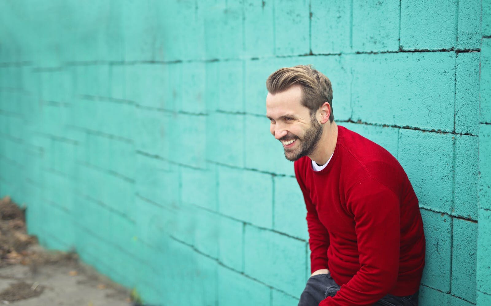 Smiling man in Budapest wearing a red sweater against a teal brick wall, exuding happiness.