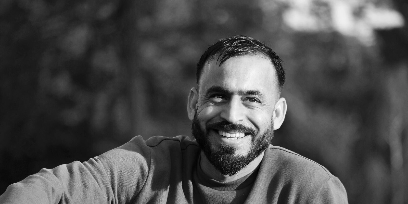 A black and white portrait of a smiling man with a beard in an outdoor setting.