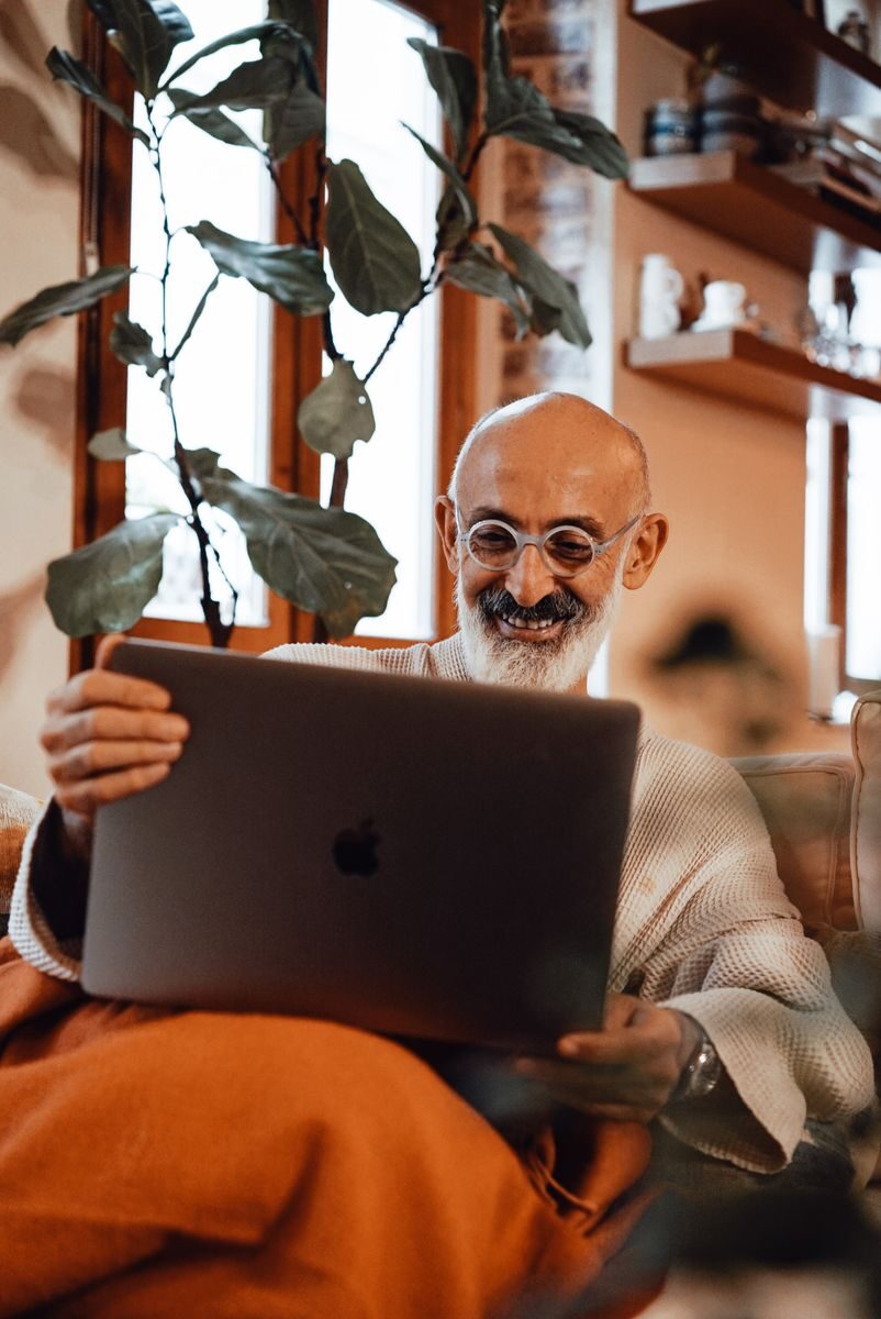 Happy bearded senior ethnic male in robe and eyeglasses resting on couch at home and smiling while having video call on laptop